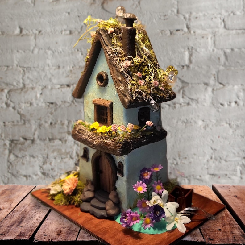 May include: A two-story light blue miniature house with a dark brown roof covered in green moss and small pink flowers. The structure features a wooden door, a balcony with yellow and pink flowers, and a base decorated with purple and white flowers.