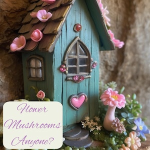 May include: A teal miniature house with a brown shingled roof adorned with pink flowers, a heart-shaped accent, and a window. A pink mushroom-shaped figure stands nearby. A white square overlay displays the text: Flower Mushrooms Anyone?