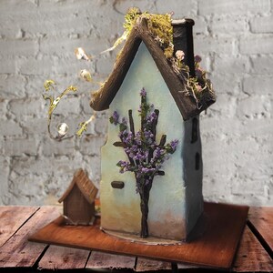 May include: A decorative miniature house with light blue walls, a dark brown roof, and a chimney. The structure features a dark brown branch-like trellis with purple flowers on the front, green moss on the roof, and a string of small warm LED lights.