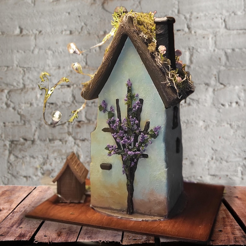 May include: A decorative miniature house with light blue walls, a dark brown roof, and a chimney. The structure features a dark brown branch-like trellis with purple flowers on the front, green moss on the roof, and a string of small warm LED lights.