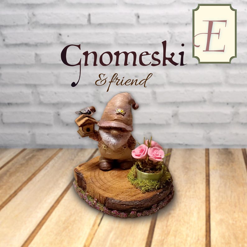 May include: A gnome figurine wearing a brown hat and overalls, holding a small birdhouse with a bird on top. Next to the gnome is a small pot with three pink roses on a circular wooden base. The text Gnomeski & friend is visible above.