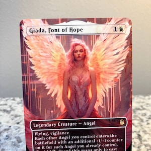 May include: A fantasy trading card featuring a woman with pink hair and white wings. The card is titled "Giada, Font of Hope" and has a power and toughness of 2/2. The card text reads: "Flying, vigilance. Each other Angel you control enters the battlefield with an additional +1/+1 counter on it for each Angel you already control. Add. Spend this mana only to cast an Angel spell."