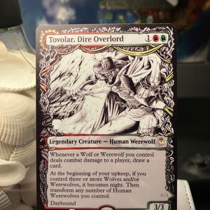 May include: A black and white illustration of a werewolf creature with a red, white, and green mana cost. The card is titled "Tovolar, Dire Overlord" and has the text "Legendary Creature - Human Werewolf". The card has the following text: "Whenever a Wolf or Werewolf you control deals combat damage to a player, draw a card. At the beginning of your upkeep, if you control three or more Wolves and/or Werewolves, it becomes night. Then transform any number of Human Werewolves you control. Daybound 4/4 3/3".