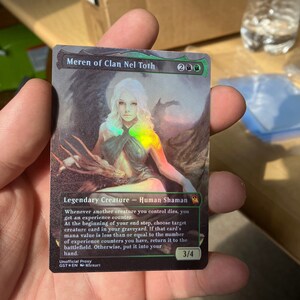 May include: A holographic trading card titled Meren of Clan Nel Toth featuring a white-haired figure in a dark setting. The card displays the text Legendary Creature - Human Shaman, a 3/4 power and toughness, and game rules text in English.