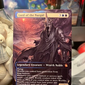 K&ouml;nnte beinhalten: Eine schwarz-wei&szlig;e Fantasy-Sammelkarte mit einer dunklen, schattenhaften Figur mit einem geh&ouml;rnten Helm. Die Karte tr&auml;gt den Titel "Lord of the Nazg&ucirc;l" und hat eine St&auml;rke und Widerstandsf&auml;higkeit von 4/3. Der Kartentext beschreibt die F&auml;higkeiten der Kreatur, darunter Fliegen und das Erschaffen von Wraith-Markern.