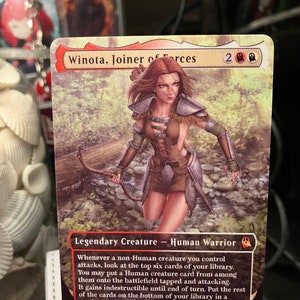 May include: A fantasy trading card featuring a woman with red hair and a bow and arrow. The card is titled "Winota, Joiner of Forces" and has a power and toughness of 4/4. The card text reads: "Whenever a non-Human creature you control attacks, look at the top six cards of your library. You may put a Human creature card from among them onto the battlefield tapped and attacking. It gains indestructible until end of turn. Put the rest of the cards on the bottom of your library in a random order."
