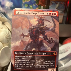 May include: A fantasy trading card featuring a red and black dragon with glowing red eyes. The card is titled "Chiss-Goria, Forge Tyrant" and has a power of 5 and a toughness of 4. The card text reads: "Legendary Creature - Dragon. Affinity for artifacts. Flying, haste. Whenever Chiss-Goria, Forge Tyrant attacks, exile the top five cards of your library. You may cast an artifact spell from among them this turn. If you do, it has affinity for artifacts."