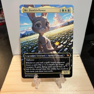 May include: A Magic: The Gathering card featuring Ms. Bumbleflower, a rabbit character, set against a vibrant field of flowers. The card includes game text and the number 1/5. The card is displayed on a clear plastic stand.