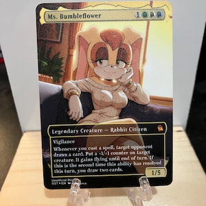 May include: A trading card featuring Ms. Bumbleflower, a rabbit character, in a cream-colored sweater. The card has a gold border and includes game text about casting spells and drawing cards. The card is propped up on a clear stand.
