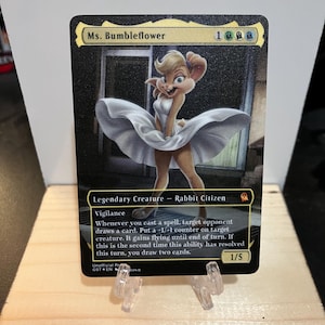 May include: A collectible card featuring Ms. Bumbleflower, a rabbit character in a white dress. The card has a black border and includes game text and a 1/5 power/toughness indicator. The card is displayed on a wooden surface.
