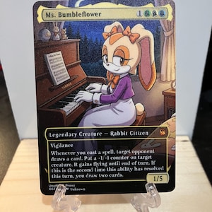 May include: A trading card featuring Ms. Bumbleflower, a rabbit character, playing a piano. The card has a night scene background with text that reads "Legendary Creature - Rabbit Citizen" and "Vigilance". The card is supported by two clear plastic stands.