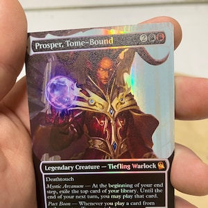 May include: A Magic: The Gathering card featuring Prosper, Tome-Bound, a legendary creature with the subtype Tiefling Warlock. The card is a holographic foil card with a black background and a colorful image of Prosper, a tiefling with horns and a dark red robe, holding a purple orb. The card text reads "Prosper, Tome-Bound", "200", "Legendary Creature - Tiefling Warlock", "Deathtouch", "Mystic Arcanum - At the beginning of your end step, exile the top card of your library. Until the end of your next turn, you may play that card.", "Pact Boon - Whenever you play a card from exile, create a Treasure token.", "1/4".