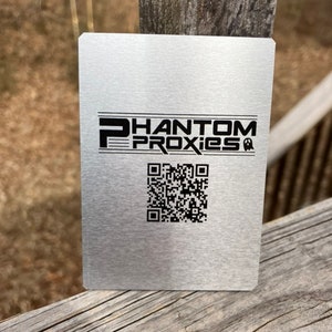 May include: A brushed metal square with the text "PHANTOM PROXIES" in black, along with a QR code. The square is resting on a weathered wooden surface.