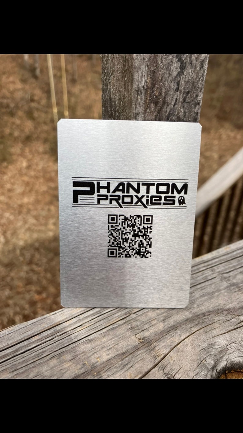 May include: A brushed metal square with the text "PHANTOM PROXIES" in black, along with a QR code. The square is resting on a weathered wooden surface.