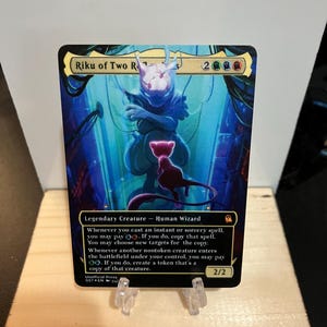 May include: A collectible card featuring the character "Riku of Two Reflections." The card has a vibrant blue and purple background with an illustration of a humanoid figure and a cat-like creature. The card text includes the creature type, abilities, and power/toughness.