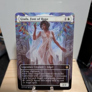 Könnte beinhalten: Eine Magic: The Gathering Sammelkarte mit Giada, Font of Hope. Die Karte zeigt eine geflügelte Figur in einem weißen Gewand mit bunten Flügeln, gekennzeichnet als legendäre Kreatur Engel mit 2/2 Stärke und Widerstandskraft.