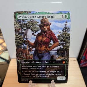 Puede incluir: Una carta coleccionable titulada Ayula, Queen Among Bears que muestra a un oso antropomórfico con un sombrero de Smokey Bear, una camisa de cuadros roja y vaqueros azules, sosteniendo una pala en un entorno forestal con ciervos.