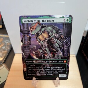Puede incluir: Una carta coleccionable con una ilustración de Michelangelo de las Tortugas Ninja con una máscara naranja. El texto de la carta dice Michelangelo, the Heart, Legendary Creature, Mutant Ninja Turtle y Raid.