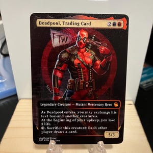 May include: A trading card featuring Deadpool, a fictional superhero, in a red and black suit. The card is black with red accents and features the text "Deadpool, Trading Card" and "Legendary Creature - Mutant Mercenary Hero". The card also has the text "As Deadpool enters, you may exchange his text box and another creature's. At the beginning of your upkeep, you lose 3 life. Sacrifice this creature: Each other player draws a card." The card has a power of 5 and a toughness of 3.