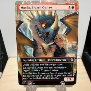 May include: A collectible trading card titled Magda, Brazen Outlaw, featuring a blue and orange dragon with a blonde character. The card text identifies it as a Legendary Creature - Dwarf Berserker with a power and toughness of 2/1 and game rules.