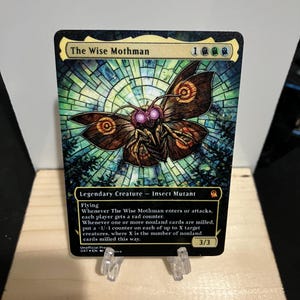 May include: A Magic: The Gathering card featuring "The Wise Mothman" with a stained-glass background. The card has a black border, with text describing the creature's abilities and stats. The card is displayed on a wooden stand.