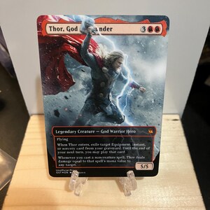 May include: A rectangular trading card featuring an illustration of Thor holding a hammer with a red cape and lightning. The text reads Thor, God of Thunder, Legendary Creature - God Warrior Hero, Flying, and includes game rules and 5/5 stats.