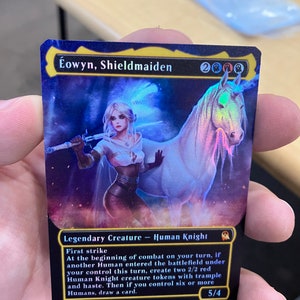 May include: A fantasy trading card featuring a woman with a sword and a unicorn. The card is black with gold trim and features the text "Éowyn, Shieldmaiden" and "Legendary Creature - Human Knight". The card has a power and toughness of 5/4.