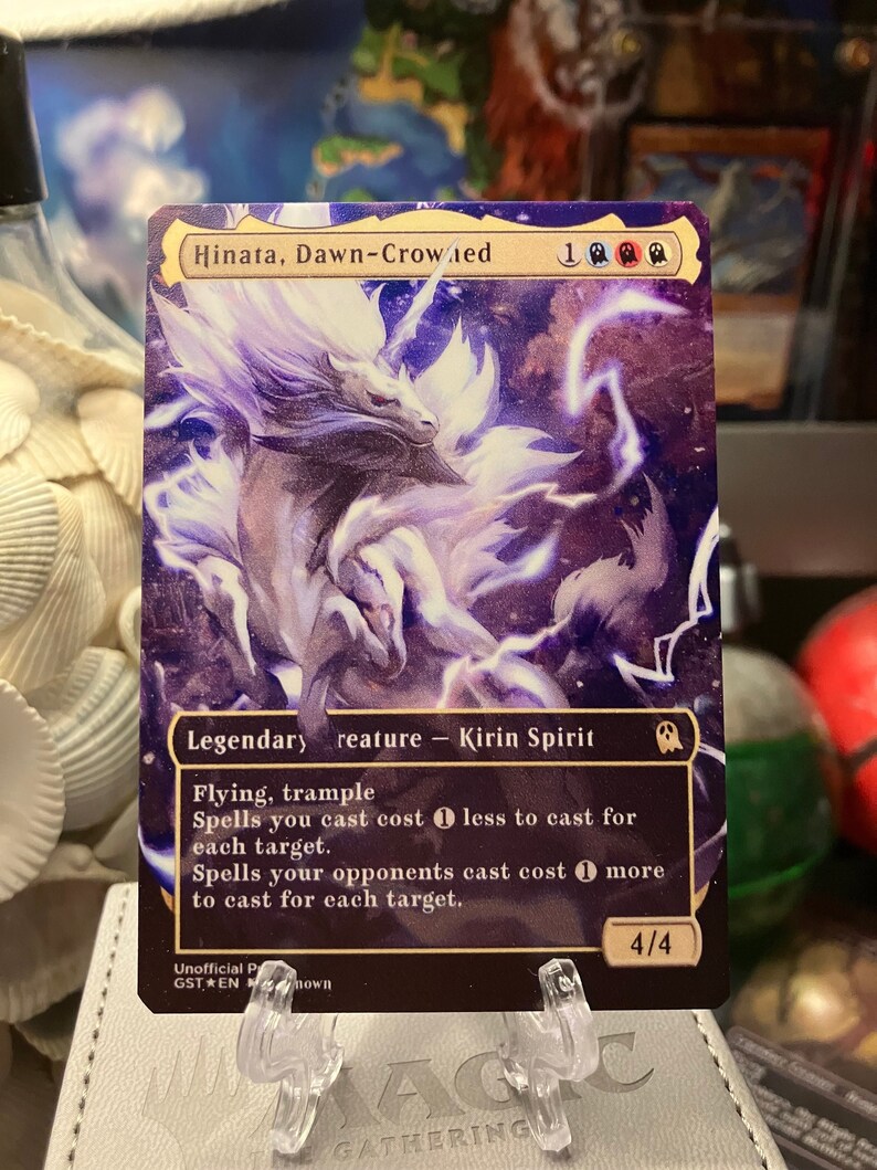 Puede incluir: Una carta de Magic: The Gathering con Hinata, Coronada del Amanecer, una criatura legendaria Esp&iacute;ritu Kirin con las habilidades de volar y arrollar. La carta tiene un fondo morado y blanco con una ilustraci&oacute;n de criatura blanca. El texto de la carta dice: "Los hechizos que lanzas cuestan 1 menos de lanzar por cada objetivo. Los hechizos que tus oponentes lanzan cuestan 1 m&aacute;s de lanzar por cada objetivo."