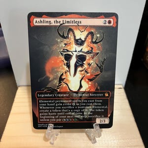 May include: A Magic: The Gathering card featuring "Ashling, the Limitless." The card has a black background with a fiery orange and white illustration of a horned figure. Text includes the card's name, creature type, and game rules. The card is propped up on a clear stand.