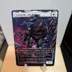Puede incluir: Una carta coleccionable rectangular que muestra un personaje de tortuga verde con máscara y caparazón azules, sosteniendo dos espadas. La carta muestra el texto Leonardo, the R-ce, Legendary Creature - Mutant Ninja Turtle y reglas con un valor 3/3.