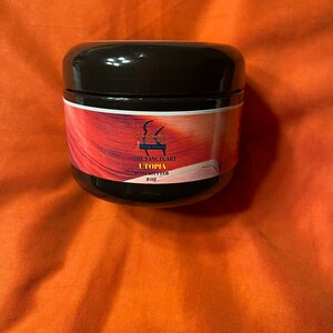 May include: A brown jar with a black lid containing Utopia body butter. The jar has a label with the text "The Sanctuary Your Temple" and "Utopia Body Butter 8 oz."