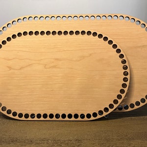May include: Two oval-shaped wooden basket bases. The bases are light brown with a series of evenly spaced holes around the perimeter. The wood grain is visible. The bases are of different sizes, with one slightly larger than the other.