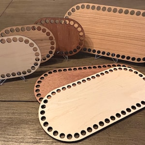 May include: A collection of oval-shaped wooden basket bases in various sizes and wood tones. Each base features a series of evenly spaced holes around the perimeter for weaving. The bases are displayed on clear stands, showcasing their different sizes and wood grains.