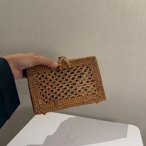 May include: A rectangular, woven rattan handbag in a natural brown hue. The bag features a lattice design on the front and a small handle at the top. The bag is held by a person.