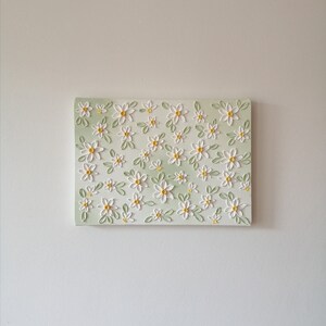 May include: A rectangular canvas art piece featuring a repeating pattern of white daisies with yellow centers and green leaves. The background has a soft, light green gradient. The artwork is mounted on a white wall.
