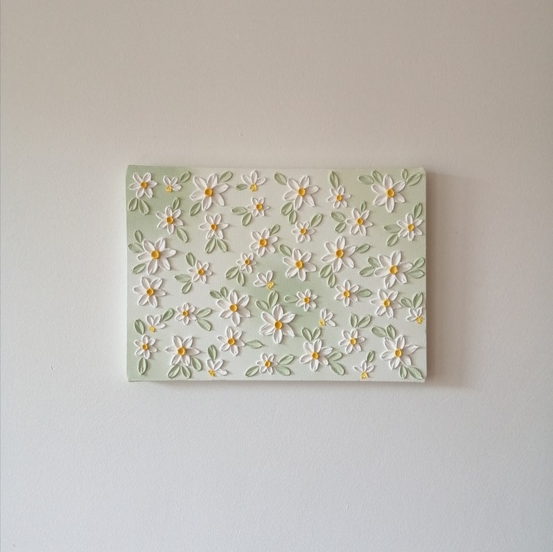 May include: A rectangular canvas art piece featuring a repeating pattern of white daisies with yellow centers and green leaves. The background has a soft, light green gradient. The artwork is mounted on a white wall.