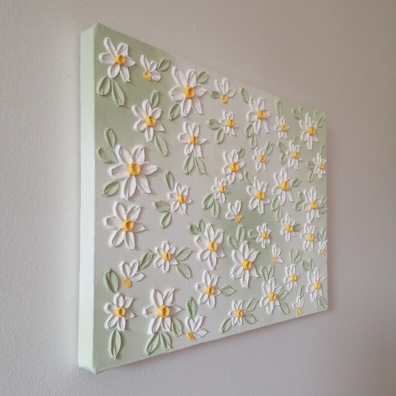 May include: A textured canvas painting featuring white daisies with yellow centers and green leaves. The flowers are arranged on a light green and white background, creating a three-dimensional effect. The canvas is mounted on a wooden frame.