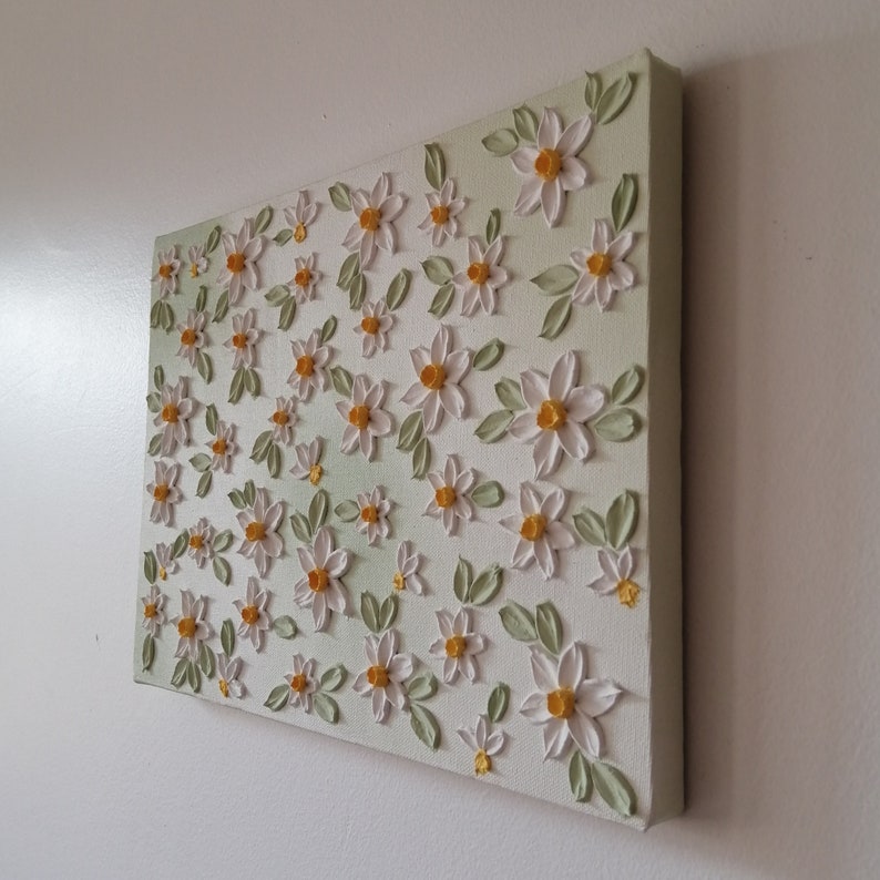 May include: A square canvas art piece featuring a textured floral design. White daisies with yellow centers and green leaves are arranged across a light green background. The artwork is mounted on a white wall.