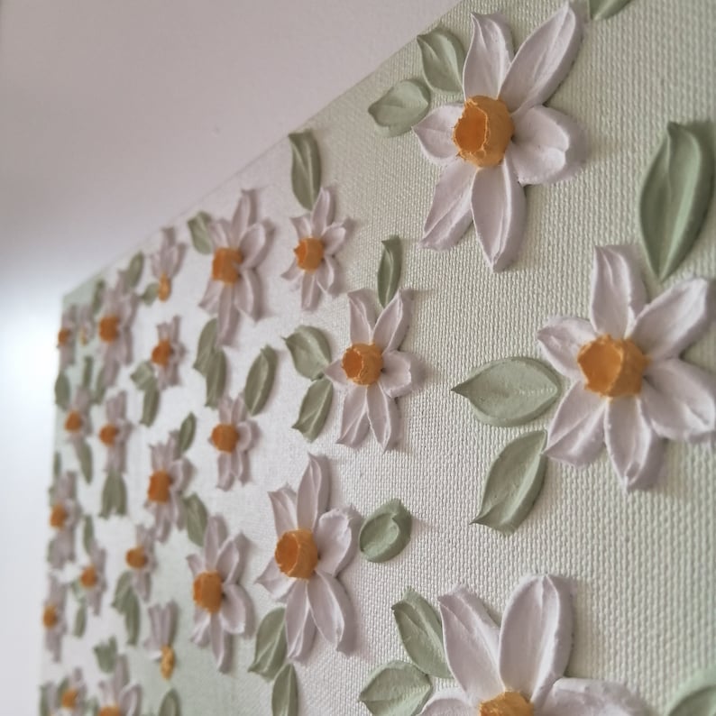 May include: A canvas art piece featuring a textured floral design. White flowers with orange centers and green leaves are arranged across a light green background. The artwork is mounted on a white frame, creating a three-dimensional effect.