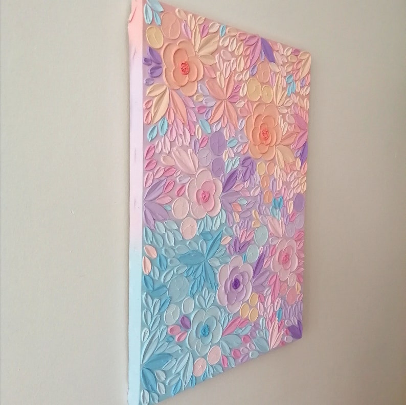 Puede incluir: Pintura en relieve floral en tonos pastel sobre lienzo. La obra presenta una superficie texturizada con flores y hojas en relieve en tonos rosa, melocot&oacute;n, azul y lavanda. El lienzo est&aacute; montado sobre un marco de madera, creando un efecto tridimensional.