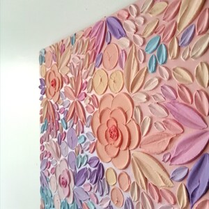 Puede incluir: Una obra de arte floral en colores pastel con texturas en relieve. El lienzo presenta una variedad de flores y hojas en tonos rosa, melocot&oacute;n, amarillo, morado y azul. La obra est&aacute; montada en una pared blanca.