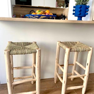 May include: Two rustic bar stools with woven seats and light-coloured wooden frames. The stools have a natural, unfinished look, with visible wood grain. The seats are square and appear to be made of woven straw or a similar material. The stools are placed in front of a kitchen counter.