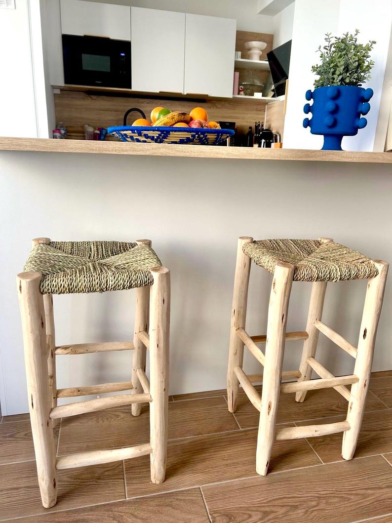 May include: Two rustic bar stools with woven seats and light-coloured wooden frames. The stools have a natural, unfinished look, with visible wood grain. The seats are square and appear to be made of woven straw or a similar material. The stools are placed in front of a kitchen counter.
