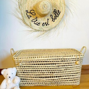 Puede incluir: Cofre de almacenamiento de paja tejida con forma rectangular y asas. Un sombrero de paja con la frase "La vie est belle" y un coraz&oacute;n rojo cuelga sobre el cofre. Un oso de peluche blanco est&aacute; sentado junto al cofre.