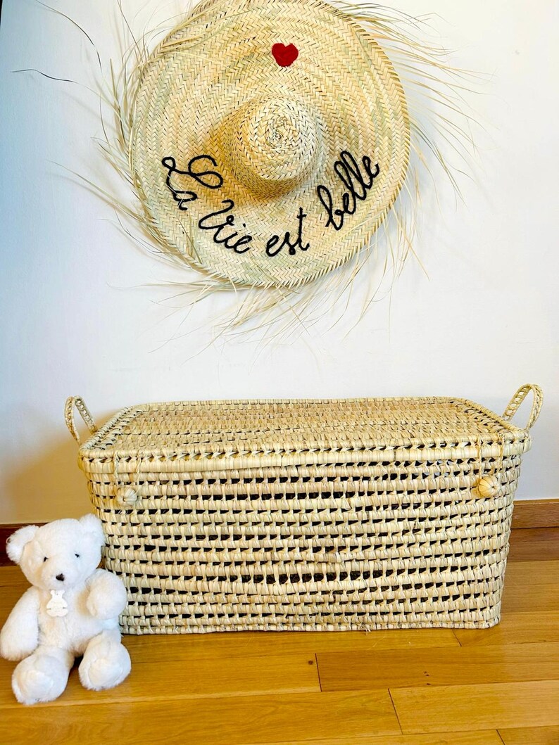 Puede incluir: Cofre de almacenamiento de paja tejida con forma rectangular y asas. Un sombrero de paja con la frase "La vie est belle" y un coraz&oacute;n rojo cuelga sobre el cofre. Un oso de peluche blanco est&aacute; sentado junto al cofre.