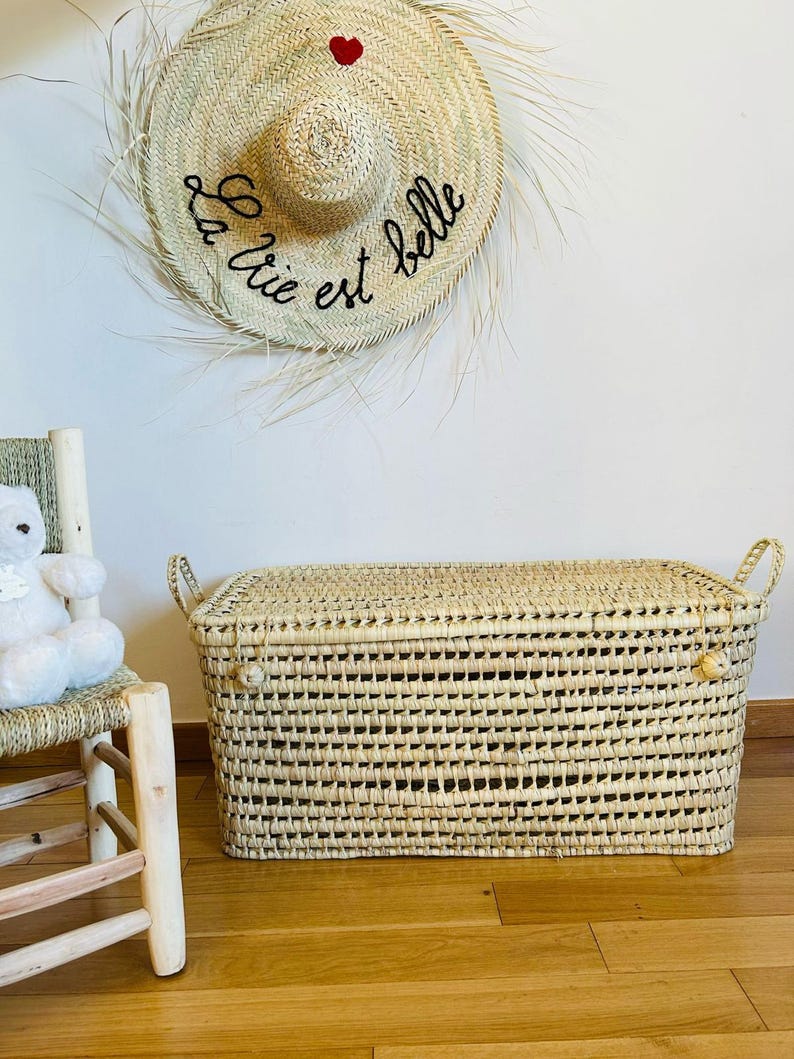 Puede incluir: Un cofre de almacenamiento rectangular tejido con tapa y asas. El cofre est&aacute; hecho de material tejido de color natural. Un sombrero de paja con el texto "La vie est belle" y un coraz&oacute;n rojo cuelga de la pared.