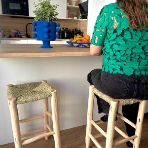 May include: Two wooden bar stools with woven seats. A blue vase with a plant sits on a wooden countertop. A person wearing a green lace shirt sits at the counter. A bowl of fruit is also on the counter.
