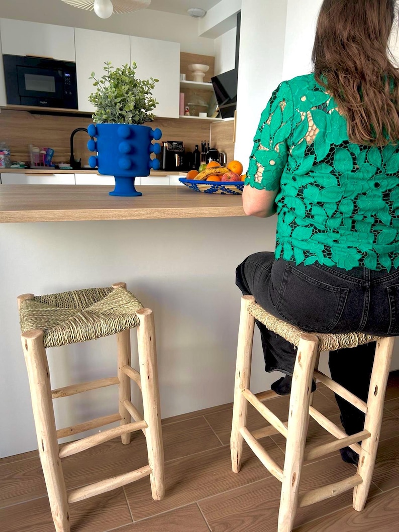 May include: Two wooden bar stools with woven seats. A blue vase with a plant sits on a wooden countertop. A person wearing a green lace shirt sits at the counter. A bowl of fruit is also on the counter.