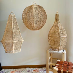 Peut inclure: Trois lampes suspendues en rotin tressé, une ronde, une conique et une en forme de vase, suspendues au plafond d'une pièce aux murs blancs et un tapis blanc avec des accents orange et violet.