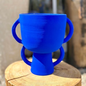 Peut inclure: Un vase décoratif bleu vif avec deux paires de poignées. Il est posé sur une tranche de bois naturelle, mettant en valeur sa couleur vive et son design unique. Le vase est de forme moderne et sculpturale, idéal pour la décoration intérieure.